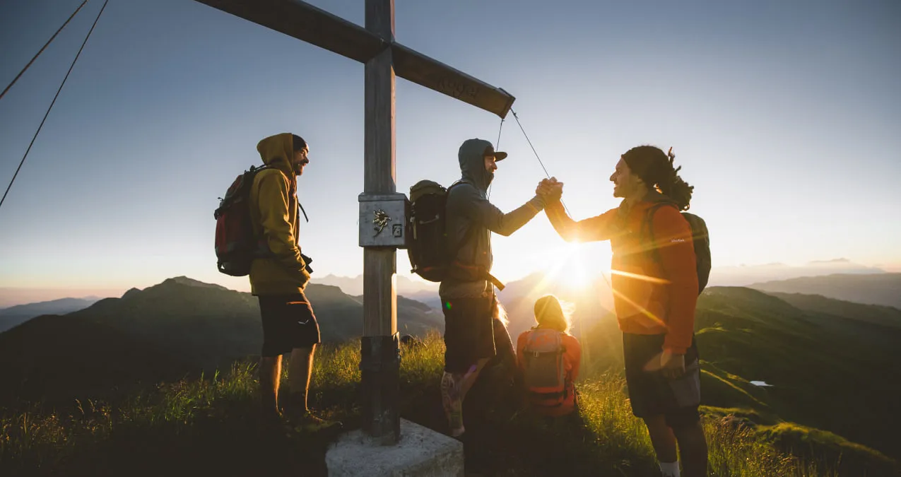 Vier Wanderer stehen am Gipfelkreuz bei Sonnenuntergang in Saalbach-Hinterglemm
