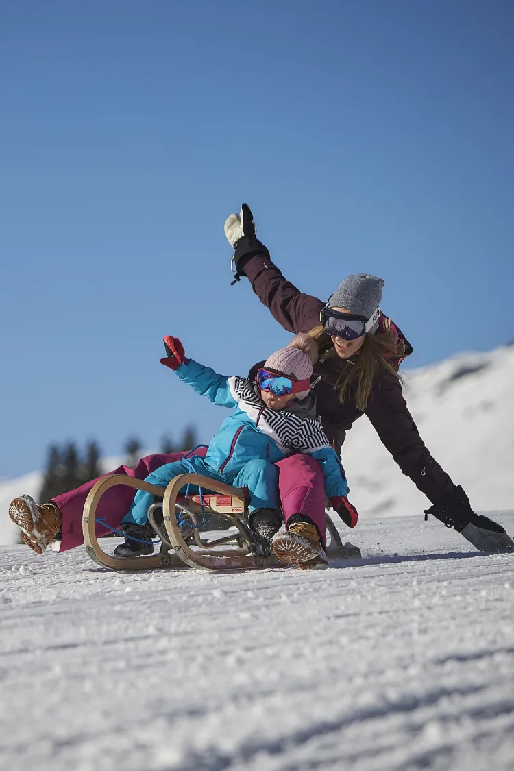 Zwei Personen rodeln auf einem verschneiten Hang in den Bergen.