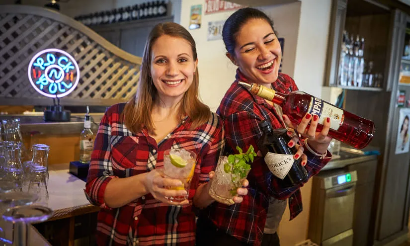 Two bartenders with cocktails behind the bar, smiling and holding drinks in hand