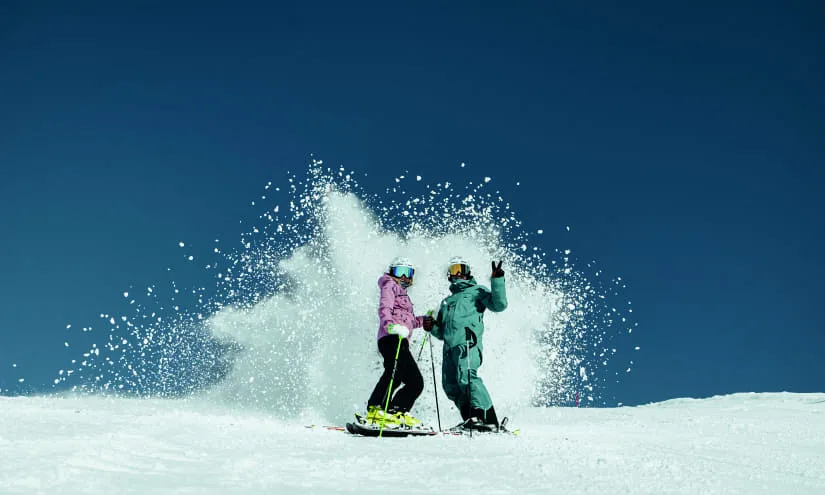 Zwei Personen ski fahren auf schneebedecktem Gelände mit einem Schneespritzer dahinter