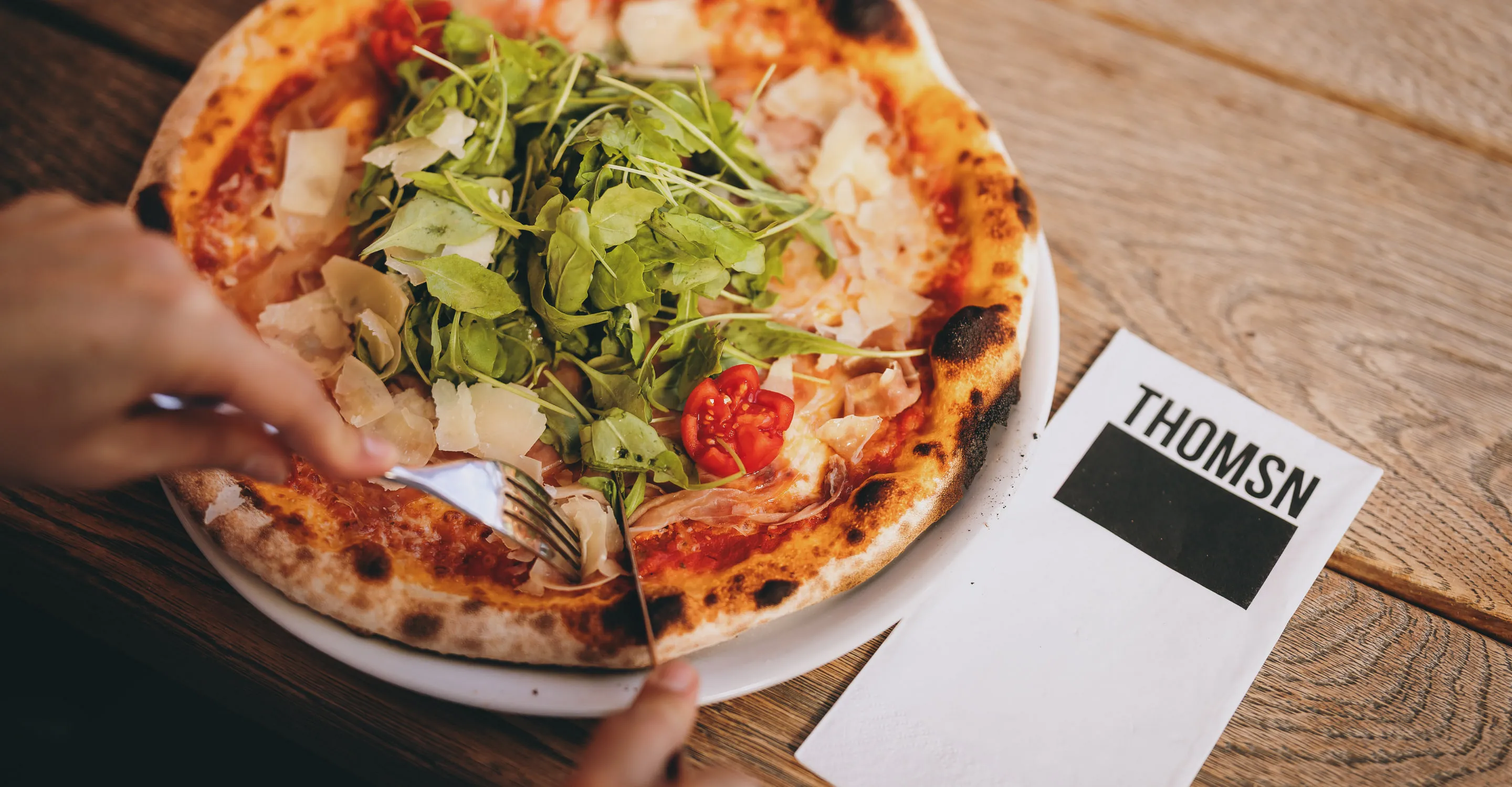 A person cuts a pizza topped with salad on a table at THOMSN.