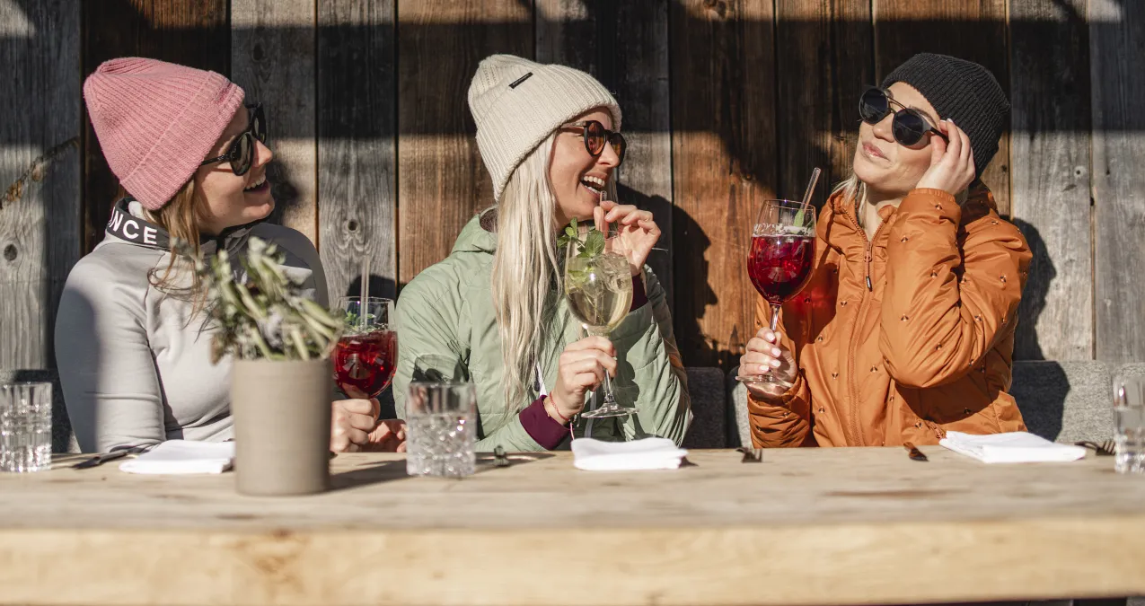 Three women with drinks sit at a table in front of a wooden wall.