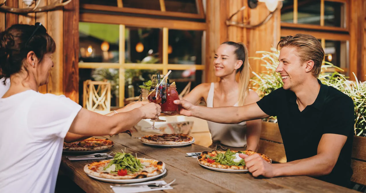 Friends toast with drinks, surrounded by pizza and food.