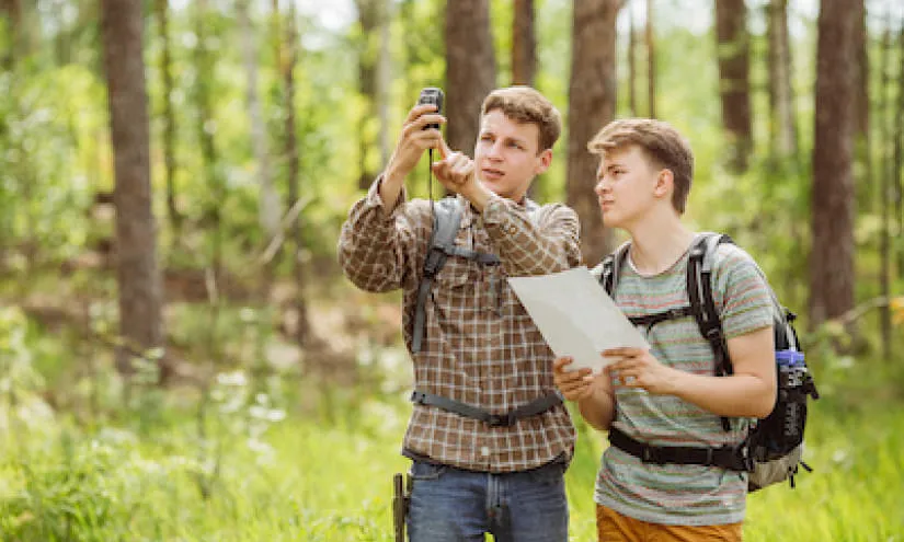 Zwei junge Männer mit Rucksäcken nutzen ein Smartphone und ein Blatt Papier im Wald.