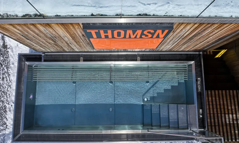Luftaufnahme eines Schwimmbeckens mit Treppen, neben einer holzverkleideten Struktur mit der Aufschrift THOMSN.