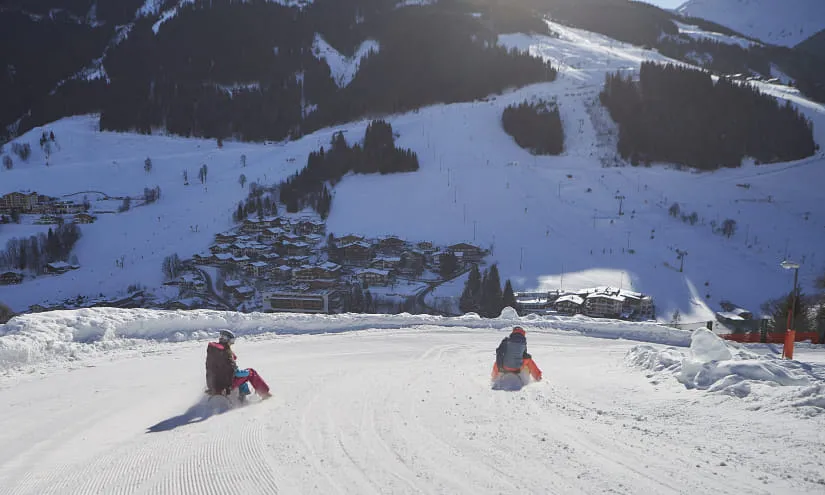 Zwei Personen rodeln auf einer Kurve im Schnee mit Berglandschaft im Hintergrund.