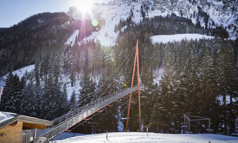 Hängeseilbrücke im Schnee, umgeben von Bergen und Wald