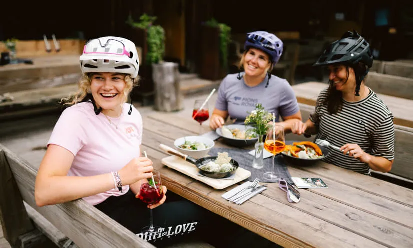 Three women in helmets enjoying food and drinks at a table.